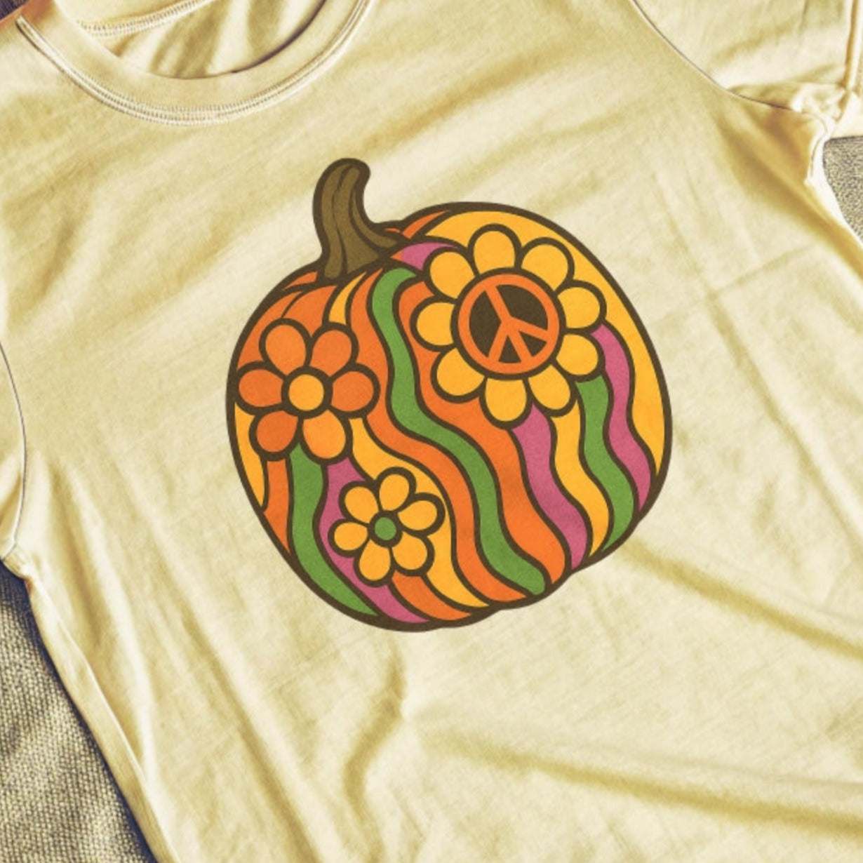 Tan t-shirt with a colorful groovy retro pumpkin design featuring peace signs and flowers, surrounded by Halloween decorations on a wooden surface.