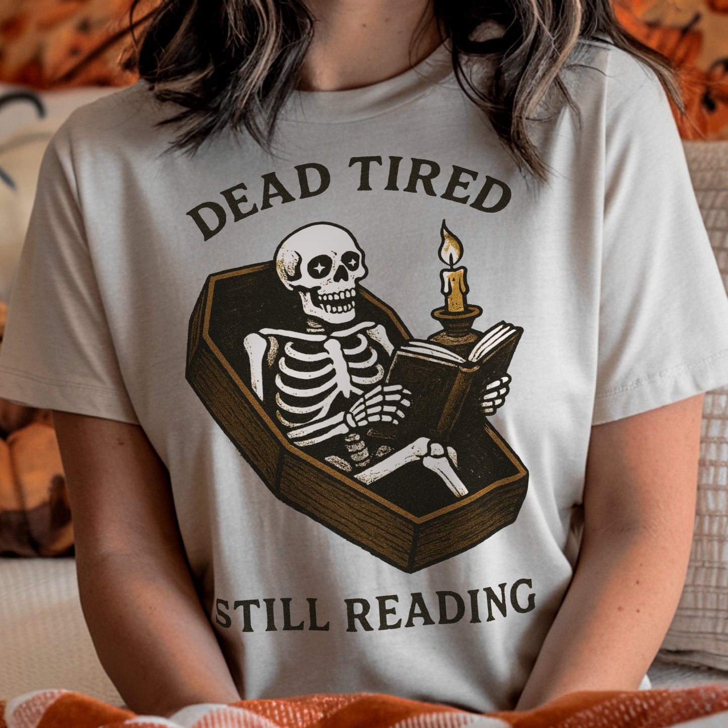 Person wearing a t-shirt with a skeleton reading a book graphic and text 'Dead Tired Still Reading'.