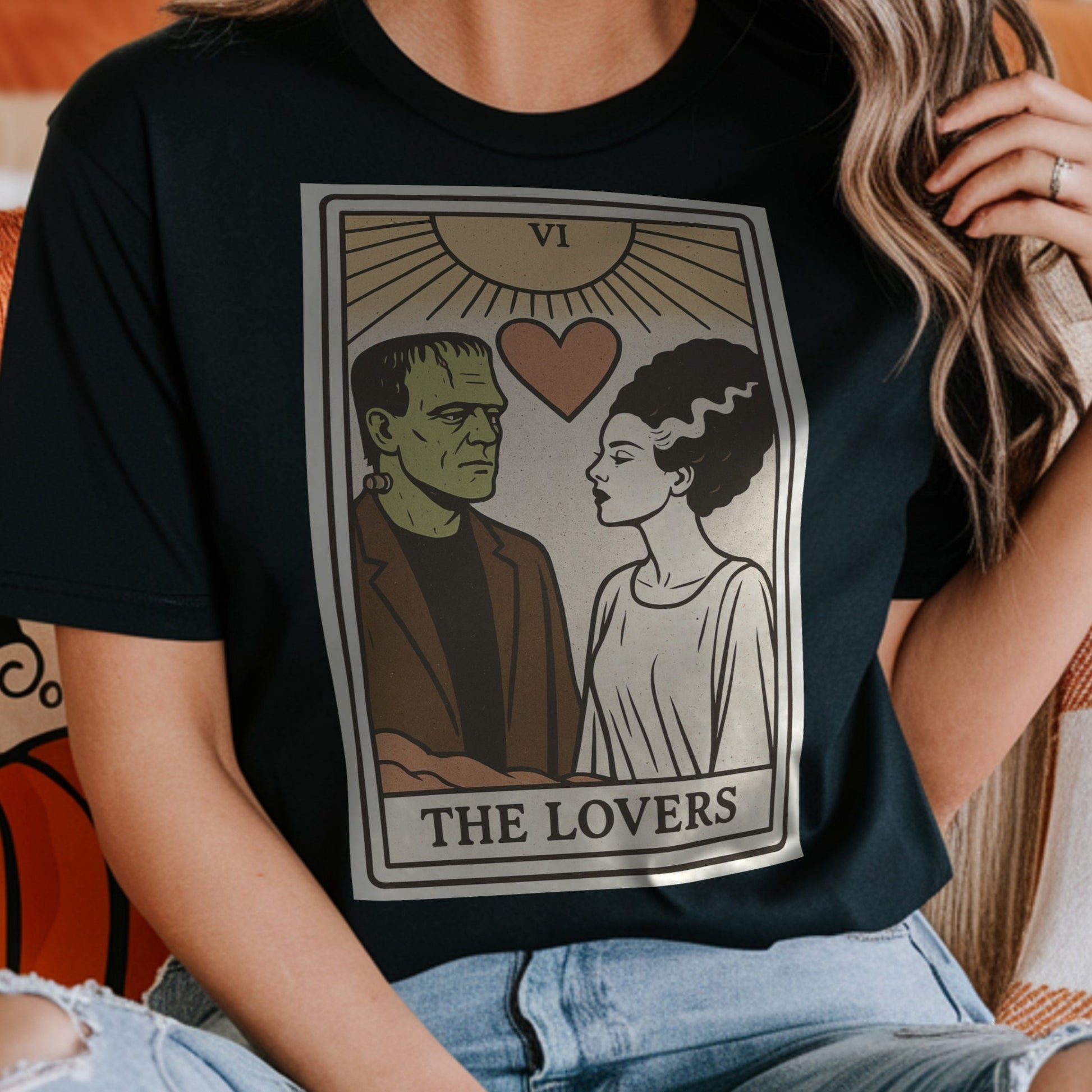 Person wearing a black t-shirt with 'The Lovers' tarot card design with Frankenstein's monster and the Bride of Frankenstein, sitting on a couch with pumpkins in the background.