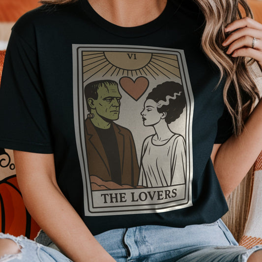 Person wearing a black t-shirt with 'The Lovers' tarot card design with Frankenstein's monster and the Bride of Frankenstein, sitting on a couch with pumpkins in the background.