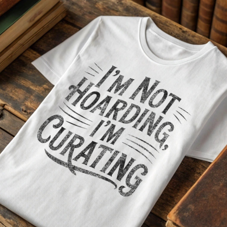 White t-shirt with 'I'm not hoarding, I'm curating' text on a wooden surface with books.