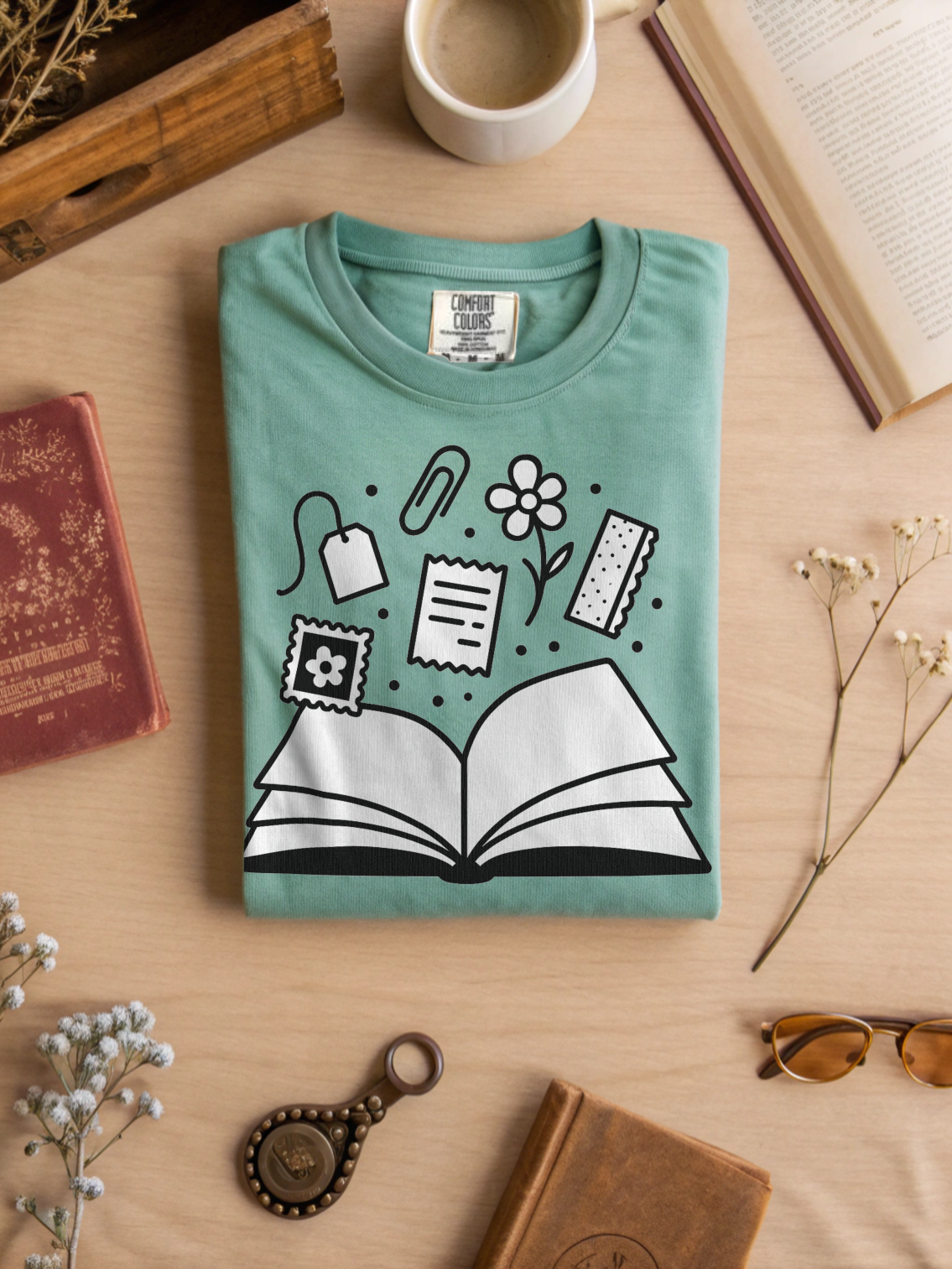 Folded teal t-shirt with book and icon design on a wooden surface with books and a cup.