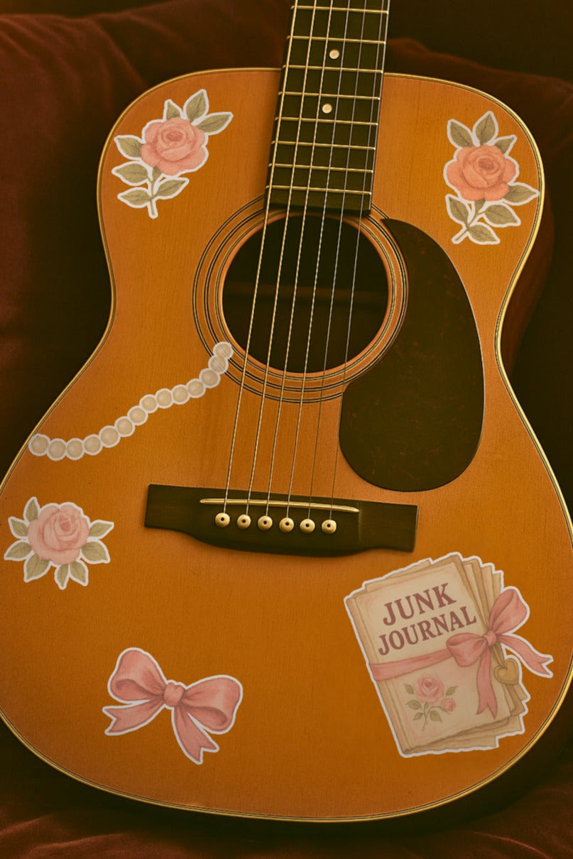 Acoustic guitar with decorative stickers from the Coquette Set. Pictured are three pink roses, a pink bow, a junk journal tied with a bow, and a length of pearls.