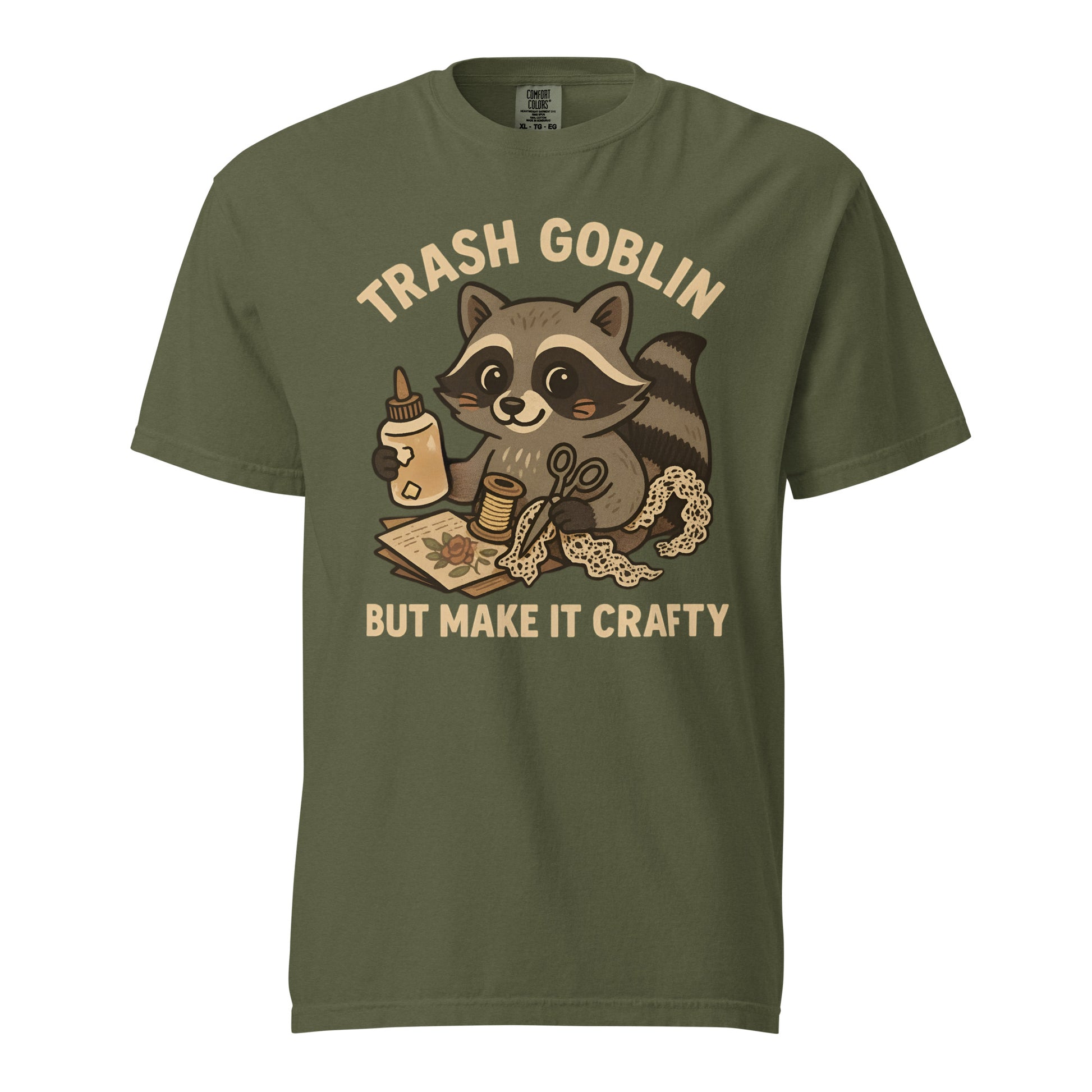 An olive green tee (color is called hemp) with a cute cartoon racoon holding craft supplies. Text says "Trash Goblin, but make it crafty."