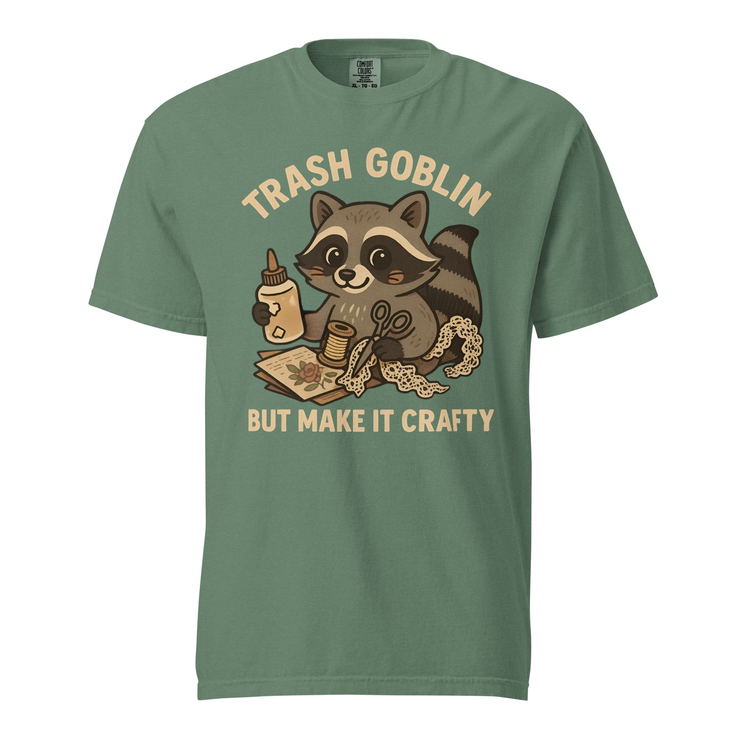 A light green tee with a cute cartoon racoon holding craft supplies. Text says "Trash Goblin, but make it crafty."