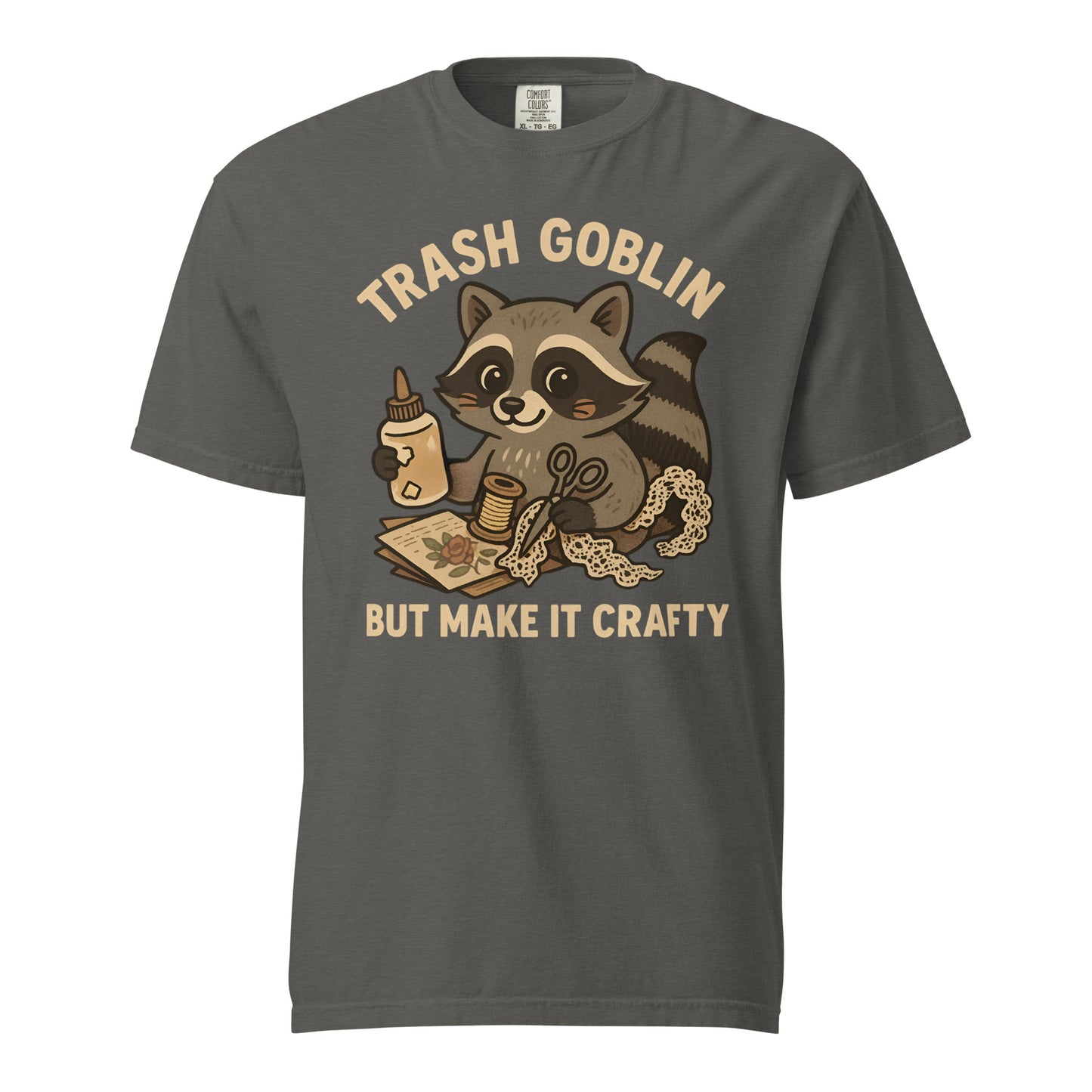 A dark grey tee (color is called Pepper) with a cute cartoon racoon holding craft supplies. Text says "Trash Goblin, but make it crafty."