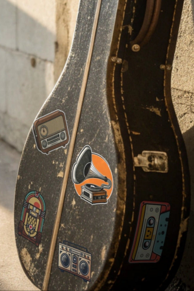 Guitar case with multiple stickers leaning against a concrete wall with sunlight casting shadows. Stickers are available individually or as a Retro Sounds Sticker set. Stickers pictured are a jukebox, a gramophone, a cassette tape, a 1980s era boombox and a 1940s era radio.