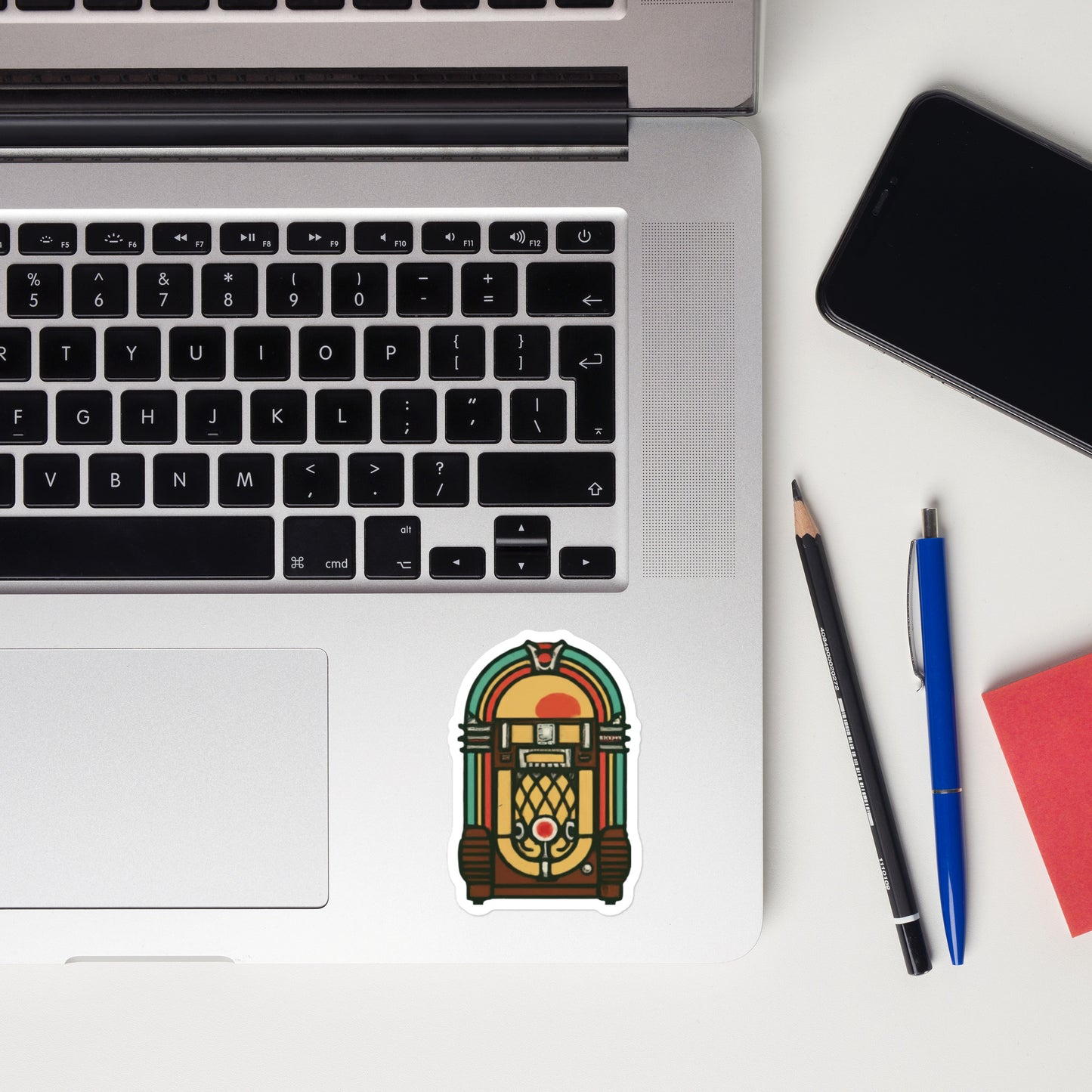 Laptop with a sticker of a vintage jukebox, next to a smartphone, pencil, pen, and notebook on a white surface.