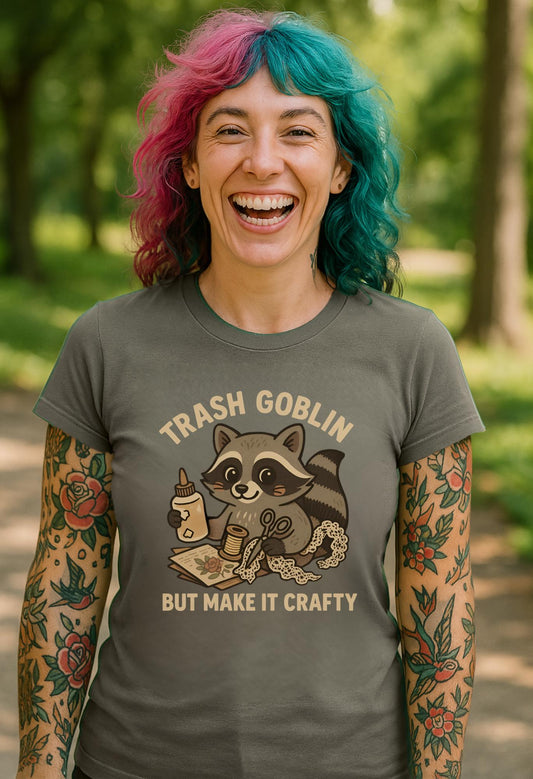 A laughing tattooed girl with pink and green hair wearing a T-shirt with a cute cartoon racoon with craft supplies. The text says "Trash Goblin but make it crafty."
